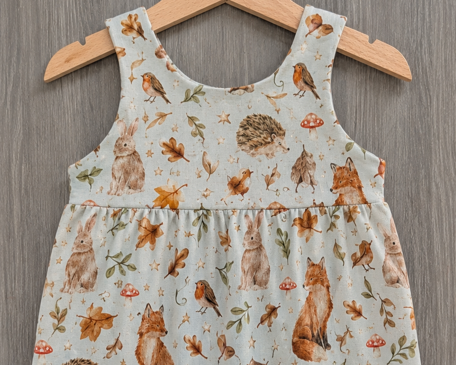 A baby sleeveless jumpsuit with an all-over woodland animal print, hanging on a wooden hanger against a gray wood-textured background. The print features foxes, rabbits, hedgehogs, and leaves on a light sage green base.